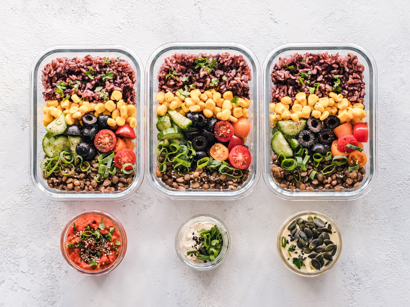 Three glass meal prep containers with rice, corn, lentils, and fresh vegetables
